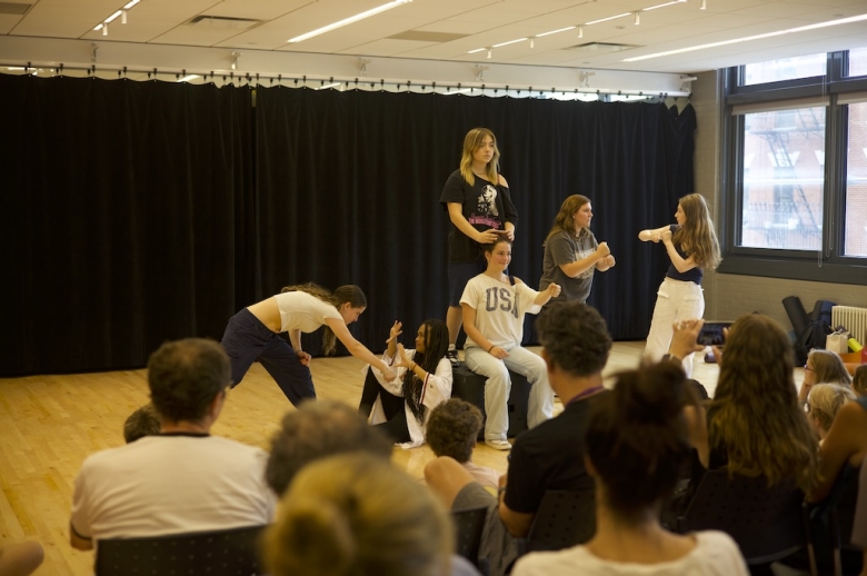 A group of Tisch Summer HIgh School Drama students pose on a stage during their final presentations on the last day of class. One student stands on a raised box while another student sits on the box in front of her. Other students pose in mid-action on either side of them.